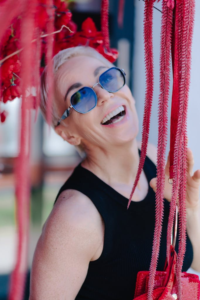 Smiling woman with blue sunglasses and vibrant red flowers enjoying a sunny day.